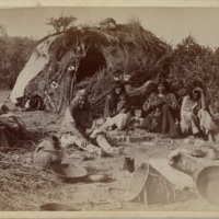 Family seated on ground outside of grass covered dwelling